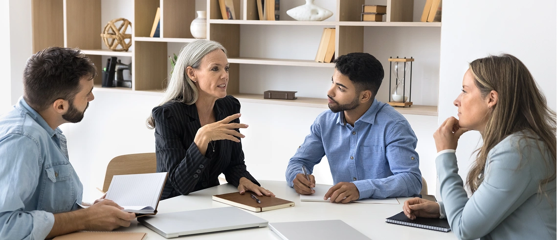 Four people sit around a table in an office, engaged in a discussion with notebooks and laptops, with shelves and decor in the background.